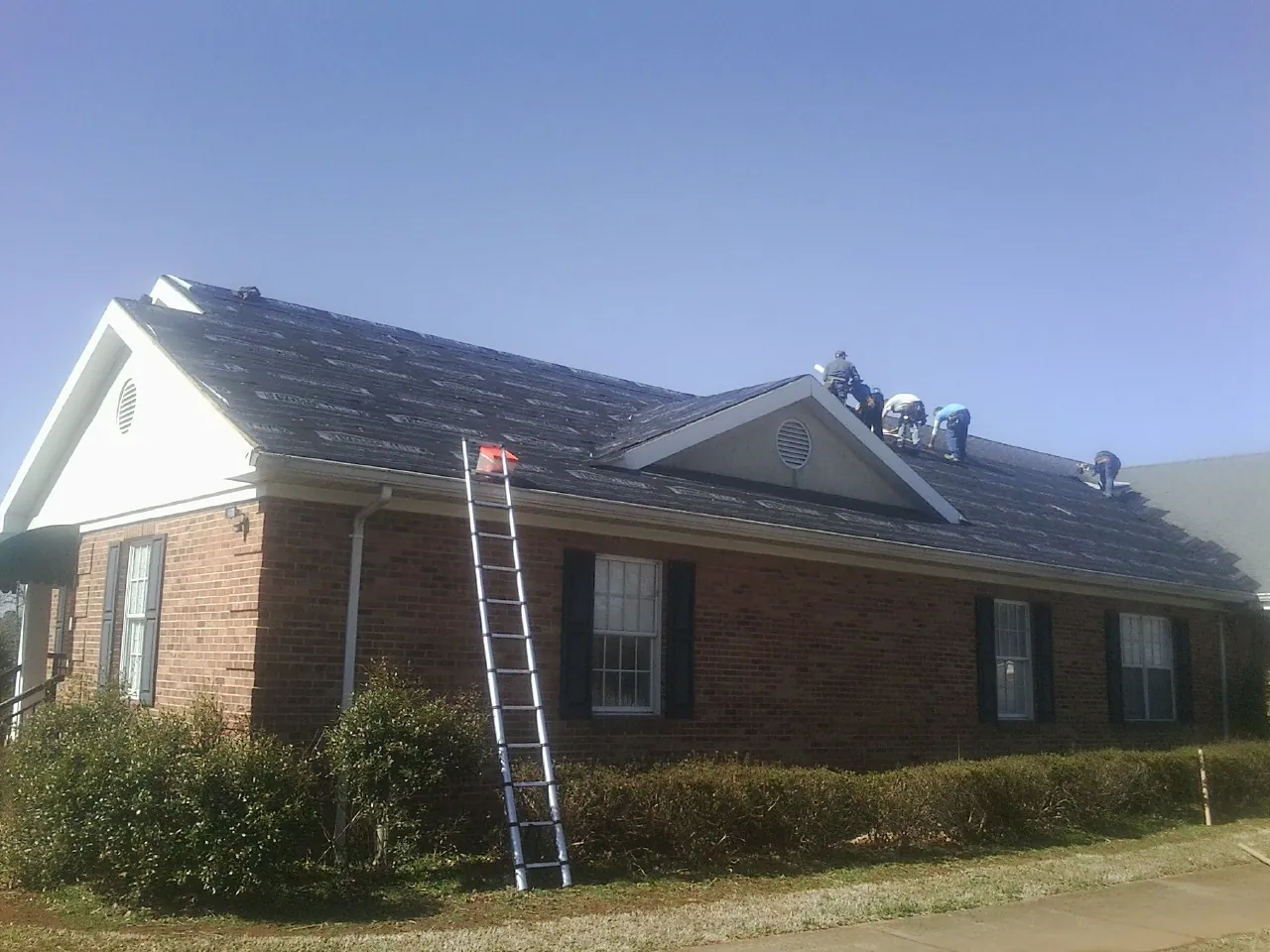 Crew tearing shingles off a brick ranch roof with a ladder set — Metal Roof Repair work by our crew in Brattleboro