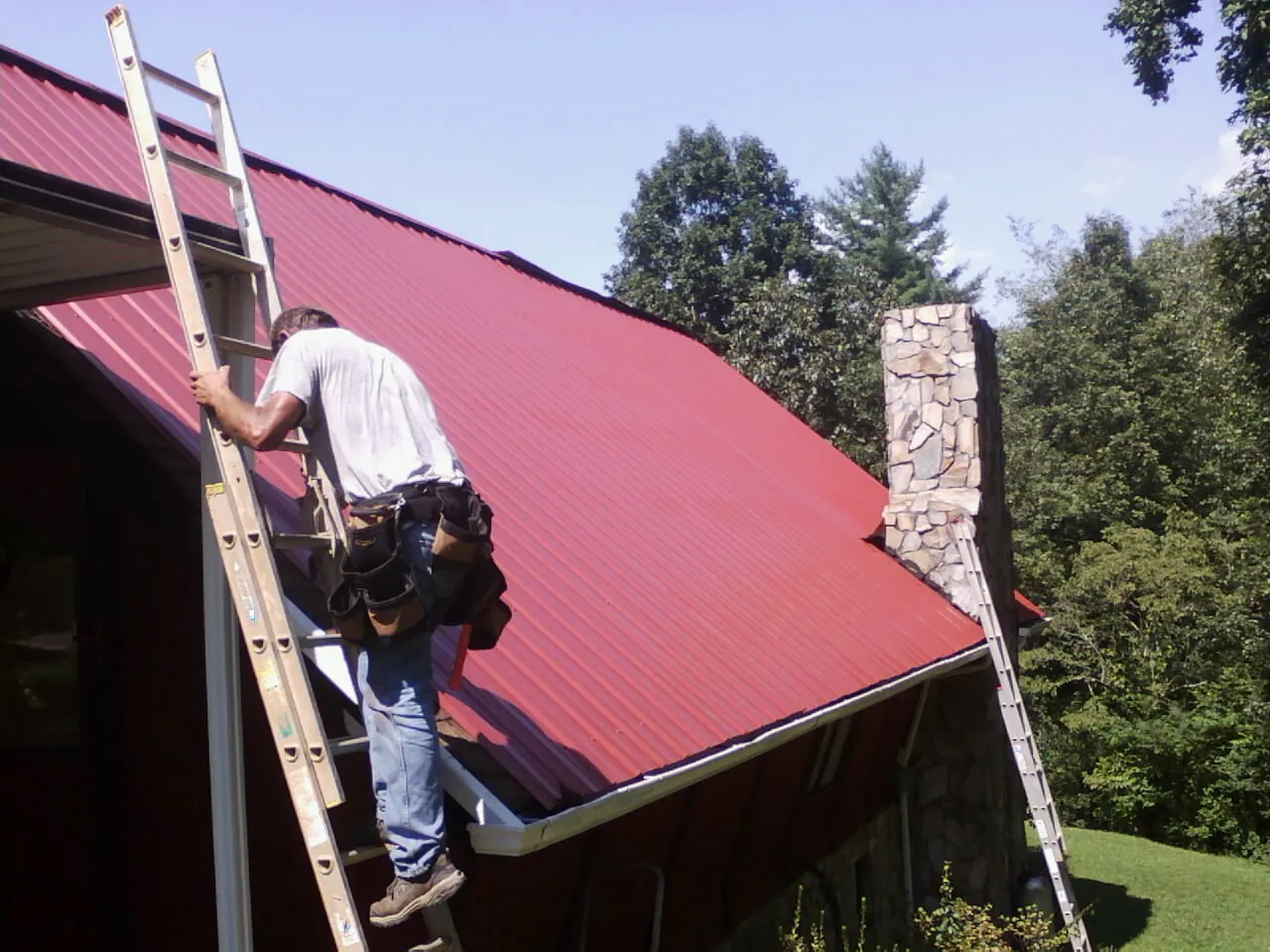 Roofer on a ladder beside a red metal roof and stone chimney — Soffit & Fascia Repair by Roofing Brothers in Brattleboro